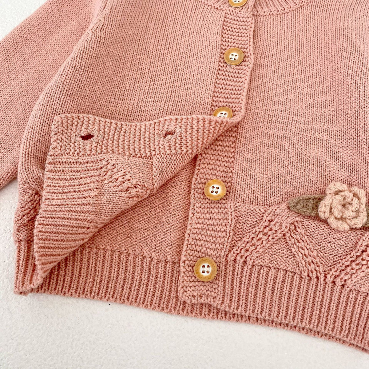 パールピンクのニットロンパース裾。花刺繍モチーフと前開きボタンのディテール