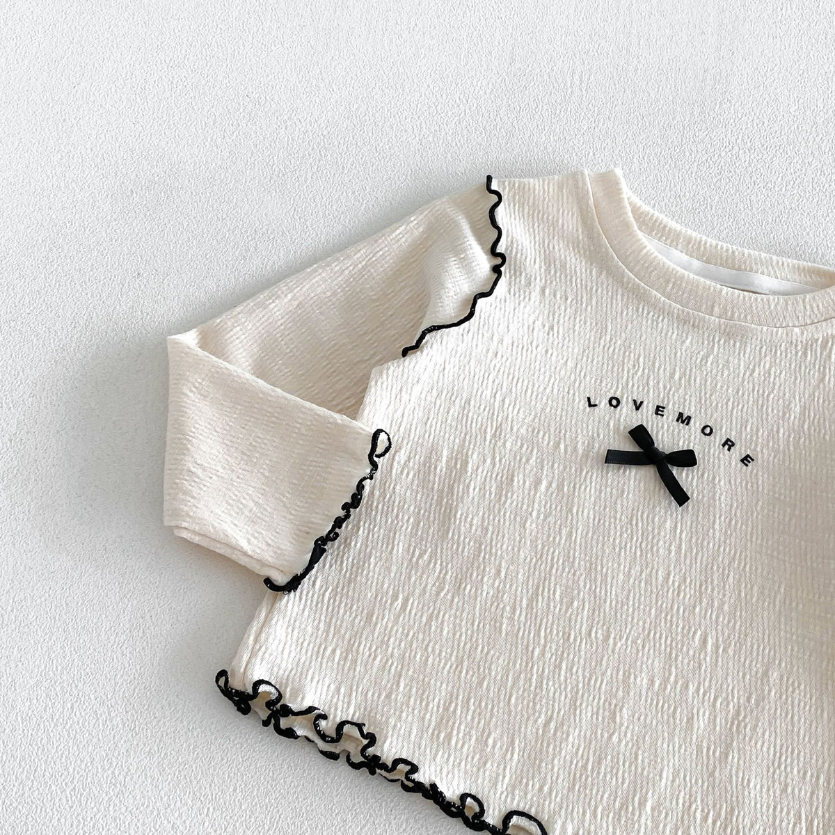 ベージュのキッズ長袖Tシャツ、ロゴと黒リボン、黒いメローフリルのライン入り